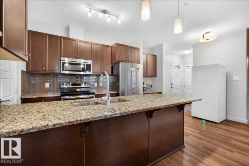 Kitchen featuring a central island with a granite-style countertop and integrated sink, rich wood-finish cabinetry, stainless steel appliances, and wood-finish flooring - 106 320 Ambleside Link Sw, Edmonton, AB - Indoor Photo Showing Kitchen With Upgraded Kitchen