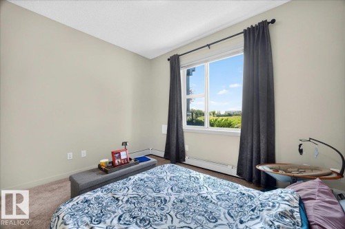 Expansive picture window with exterior scenery, neutral wall tones, textured carpeting, and white baseboards - 106 320 Ambleside Link Sw, Edmonton, AB - Indoor Photo Showing Bedroom