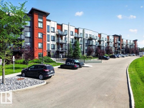 Contemporary multi-story building featuring red and grey facade elements, multiple balconies with glass railings, surrounding green landscaping, and an asphalt parking area - 106 320 Ambleside Link Sw, Edmonton, AB - Outdoor With Balcony With Facade