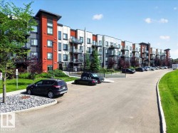 Contemporary multi-story building featuring red and grey facade elements, multiple balconies with glass railings, surrounding green landscaping, and an asphalt parking area - 