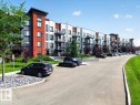 Contemporary multi-story building featuring red and grey facade elements, multiple balconies with glass railings, surrounding green landscaping, and an asphalt parking area - 106 320 Ambleside Link Sw, Edmonton, AB  - Outdoor With Balcony With Facade 