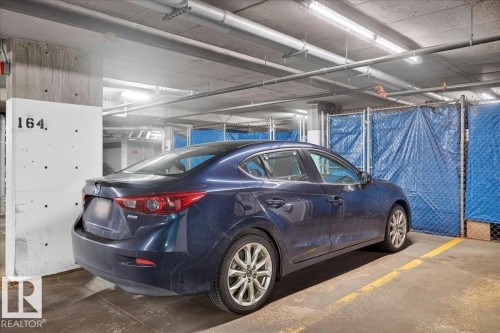 Designated parking space featuring concrete walls and ceiling, overhead lighting, and an adjacent storage unit with chain-link fencing and blue tarp enclosure - 106 320 Ambleside Link Sw, Edmonton, AB - Indoor Photo Showing Garage