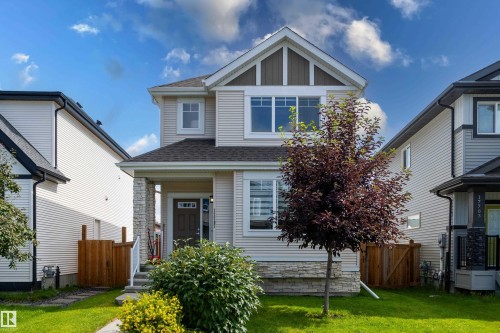 Two-story residence featuring light-colored siding, a covered front porch with stone accents, a dark brown front door, a landscaped front yard with a tree and shrubs, and a paved walkway - 17707 59 Street Nw, Edmonton, AB - Outdoor