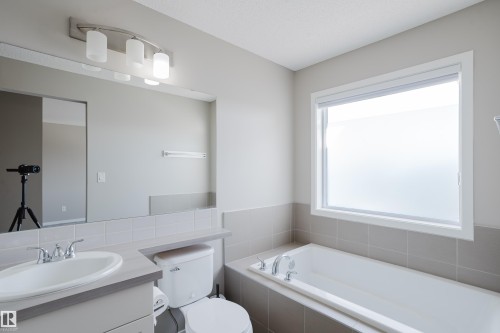 Bathroom featuring a built-in tub with tile surround, a large window, a vanity with an integrated sink, a wide mirror, and a three-light fixture - 17707 59 Street Nw, Edmonton, AB - Indoor Photo Showing Bathroom
