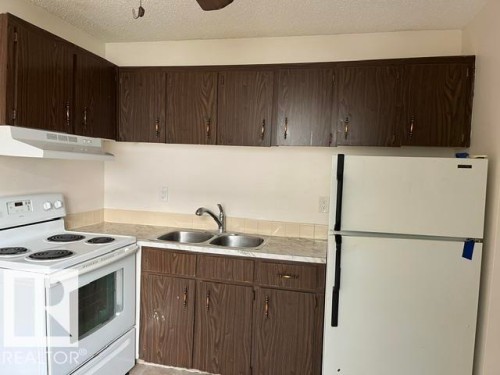 Kitchen featuring wood-finish upper and lower cabinetry, white appliances, a double basin stainless steel sink, and light-toned countertops with a tiled backsplash - #205 10740 105 St Nw, Edmonton, AB - Indoor Photo Showing Kitchen With Double Sink