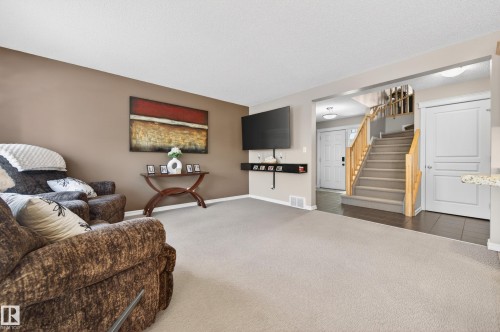 Expansive living room featuring light-toned carpet, a neutral accent wall, a built-in media shelf, and an open entryway to a staircase with wood railings - 9 Snowbird Crescent, Leduc, AB - Indoor