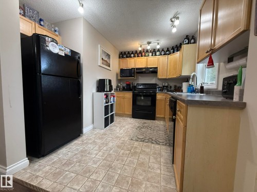 Kitchen featuring natural wood cabinetry, black appliances, track lighting, a double basin sink with a gooseneck faucet, and tile-patterned flooring - #93 12050 17 Av Sw, Edmonton, AB - Indoor Photo Showing Kitchen