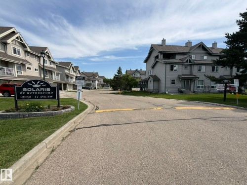 Residential community entrance featuring an illuminated sign, paved road, and a row of townhomes with balconies - #93 12050 17 Av Sw, Edmonton, AB - Outdoor With Facade
