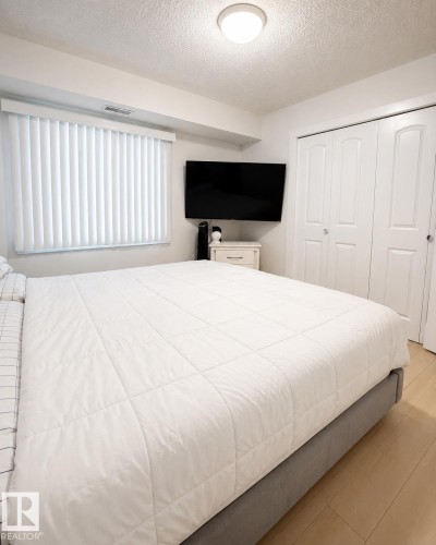 Bright bedroom featuring a large window with vertical blinds, light wood-finish flooring, a flush-mount ceiling light, and white bi-fold closet doors - #101 11825 71 St Nw, Edmonton, AB - Indoor Photo Showing Bedroom