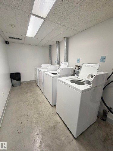 Utility room featuring three white top-loading washing machines, concrete flooring, white walls, and a suspended acoustic tile ceiling with fluorescent lighting - #101 11825 71 St Nw, Edmonton, AB - Indoor Photo Showing Laundry Room