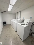 Utility room featuring three white top-loading washing machines, concrete flooring, white walls, and a suspended acoustic tile ceiling with fluorescent lighting - 