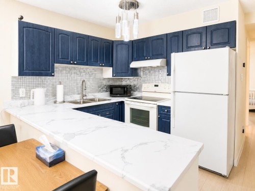 U-shaped kitchen featuring blue cabinetry, white marbled countertops, and a mosaic tile backsplash - #101 11825 71 St Nw, Edmonton, AB - Indoor Photo Showing Kitchen With Double Sink