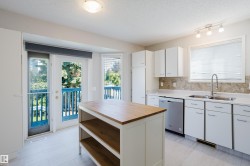 Light-filled kitchen featuring a central island with a wood-finish countertop, white cabinetry with silver hardware, a stainless steel dishwasher, and a double-basin sink with a gooseneck faucet - 