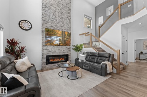 Two-story living area featuring a stacked stone fireplace with an electric insert, wood-finish flooring, and a glass-paneled staircase with natural wood handrails - 59 Willow Link, Fort Saskatchewan, AB - Indoor Photo Showing Living Room With Fireplace