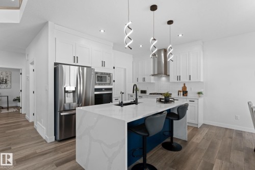 Modern kitchen featuring white shaker cabinetry, stainless steel appliances, a waterfall island with a blue base, quartz countertops, and wood-finish flooring - 59 Willow Link, Fort Saskatchewan, AB - Indoor Photo Showing Kitchen With Upgraded Kitchen