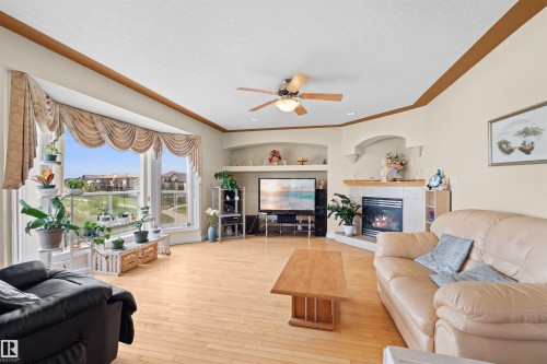 Spacious living area featuring light wood-finish flooring, a prominent bay window, a built-in corner fireplace with a tile surround and wood mantel, a ceiling fan with integrated lighting, and built-in arched display niches - 7622 168A Avenue, Edmonton, AB - Indoor Photo Showing Living Room With Fireplace