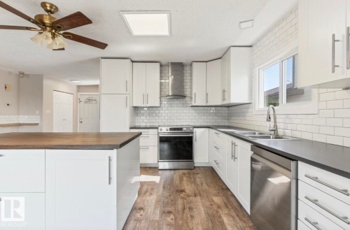 Modern kitchen featuring white cabinetry, dark countertops, a stainless steel appliance suite, subway tile backsplash, and wood-finish flooring - 10211 95 Ave, Morinville, AB - Indoor Photo Showing Kitchen With Stainless Steel Kitchen With Double Sink With Upgraded Kitchen