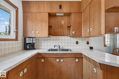 Kitchen featuring wood-finish cabinetry with decorative knobs, white patterned tile backsplash, and a double basin stainless steel sink - 12908 104 Street Nw, Edmonton, AB - Indoor Photo Showing Kitchen With Double Sink