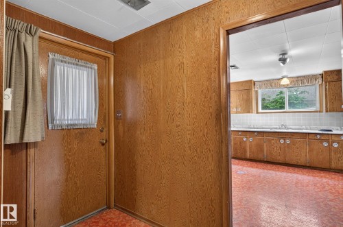 Wood-panel walls and orange-patterned flooring frame a doorway opening to a kitchen featuring wood cabinetry, a white tile backsplash, and a window above the sink - 12908 104 Street Nw, Edmonton, AB - Indoor