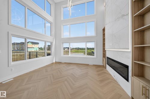 Two-story living area featuring extensive window walls, herringbone wood-finish flooring, a full-height tiled fireplace surround with an electric insert, and built-in shelving with cabinetry - 2808 191 Street Nw, Edmonton, AB - Indoor Photo Showing Other Room With Fireplace