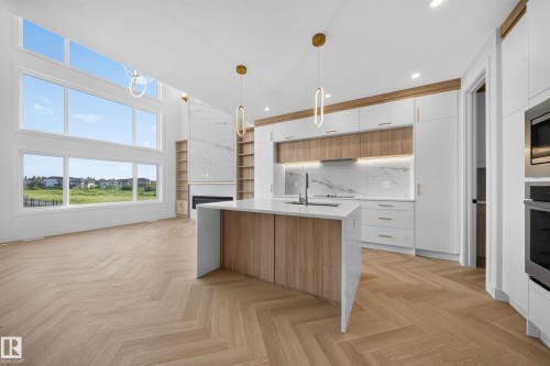 Gourmet kitchen and living area featuring high ceilings with expansive windows, wood-finish herringbone flooring, a waterfall island, marble-patterned backsplash, and a modern fireplace with built-in shelving - 2808 191 Street Nw, Edmonton, AB - Indoor Photo Showing Kitchen