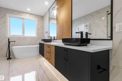 Contemporary bathroom featuring a freestanding tub with matte black floor-mounted faucet, a dual vanity with rectangular vessel sinks, dark cabinetry with wood-finish drawer fronts, and large format white tile flooring and walls - 2808 191 Street Nw, Edmonton, AB - Indoor Photo Showing Bathroom