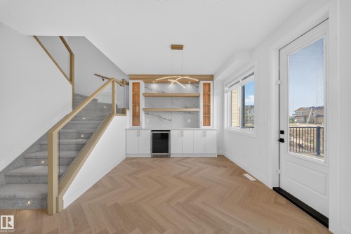 Open-concept entry area featuring herringbone wood-finish flooring, a contemporary staircase with glass railing and integrated step lighting, a built-in wet bar with a beverage cooler and display shelving, white cabinetry, and a full-light entry door - 2808 191 Street Nw, Edmonton, AB - Indoor Photo Showing Other Room
