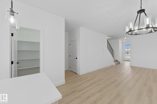 Open-concept living area featuring light wood-finish flooring, a modern chandelier, a single pendant light, a pantry with ample shelving, and a prominent staircase with a dark handrail - 4215 63 Street, Beaumont, AB - Indoor Photo Showing Other Room