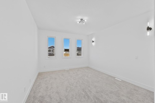 Spacious room featuring three vertical windows, plush gray carpet, a contemporary ceiling light fixture, and two wall-mounted sconces - 4215 63 Street, Beaumont, AB - Indoor Photo Showing Other Room