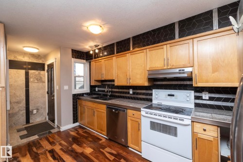 Kitchen featuring wood-finish flooring, light wood cabinetry, white electric range, stainless steel dishwasher, and dark geometric tile backsplash - 11915 77 Street Nw, Edmonton, AB - Indoor Photo Showing Kitchen