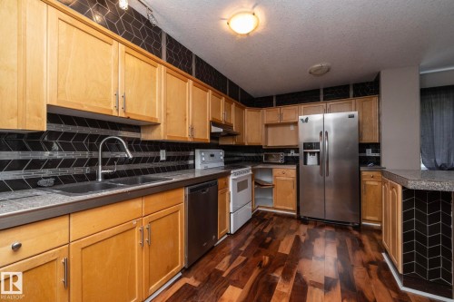 Kitchen featuring wood-finish flooring, light wood cabinetry, stainless steel appliances, a double basin sink, and a black and gray geometric tile backsplash - 11915 77 Street Nw, Edmonton, AB - Indoor Photo Showing Kitchen With Double Sink
