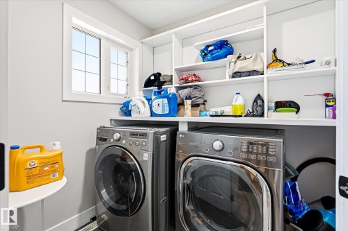 Dedicated laundry area featuring front-loading washer and dryer units, white built-in shelving, a natural light window, and a wall-mounted corner shelf - 4317 152 Avenue Nw, Edmonton, AB - Indoor Photo Showing Laundry Room