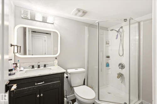 Bathroom featuring a corner glass-enclosed shower, a vanity with a white countertop and dark cabinetry, a wall-mounted mirror with integrated lighting, and a brushed nickel faucet - 4317 152 Avenue Nw, Edmonton, AB - Indoor Photo Showing Bathroom