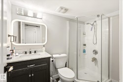 Bathroom featuring a corner glass-enclosed shower, a vanity with a white countertop and dark cabinetry, a wall-mounted mirror with integrated lighting, and a brushed nickel faucet - 