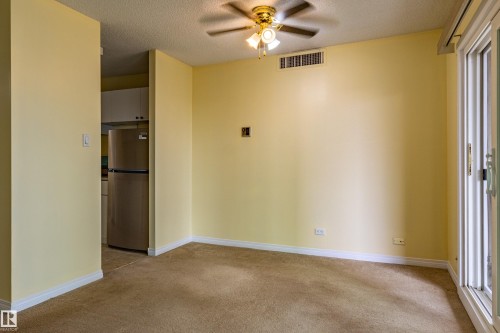 Carpeted living space featuring a ceiling fan with integrated lighting, a wall-mounted air vent, and light-toned walls - #1210 10140 120 St Nw, Edmonton, AB - Indoor Photo Showing Other Room