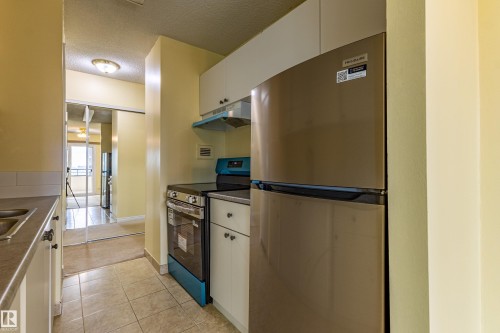 Compact kitchen featuring a stainless steel refrigerator, white cabinetry, integrated range hood, and tile flooring - #1210 10140 120 St Nw, Edmonton, AB - Indoor Photo Showing Kitchen