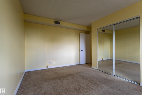 Bedroom featuring wall-to-wall carpeting, light-colored walls, white baseboards, a white interior door with dark hardware, and a mirrored closet with sliding panels - #1210 10140 120 St Nw, Edmonton, AB - Indoor Photo Showing Other Room