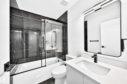 Contemporary bathroom featuring a dark marble-patterned shower surround with a sliding glass door, a white rectangular bathtub, a white vanity with a recessed sink, a rectangular mirror with rounded corners, and matte black fixtures - 