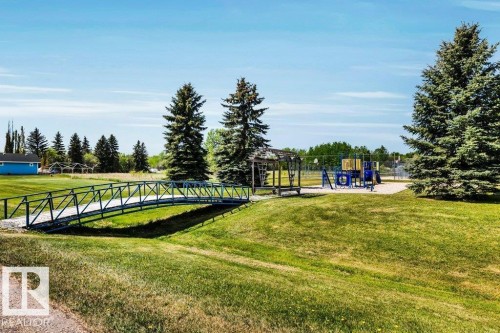 Expansive green space featuring a pedestrian bridge, mature evergreen trees, and a dedicated playground area with equipment - 12 Sunset Harbour, Rural Wetaskiwin County, AB 
