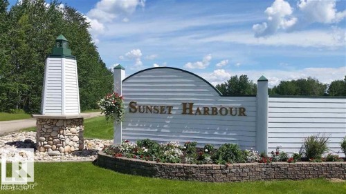 Entry sign featuring a lighthouse-style architectural element on a stone base, a decorative entry wall with horizontal siding, and a stone-edged flower bed with plantings - 12 Sunset Harbour, Rural Wetaskiwin County, AB 