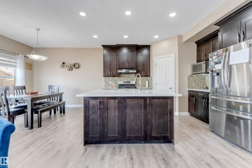 Open-concept kitchen and dining area featuring light wood-finish flooring, recessed lighting, a kitchen island with a light-colored countertop, dark wood cabinetry, and stainless steel appliances - 121 64 Street Sw, Edmonton, AB - Indoor Photo Showing Kitchen With Upgraded Kitchen