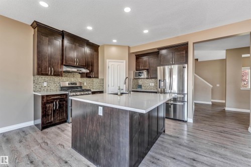 Contemporary kitchen featuring a large central island with a light-toned countertop, dark wood cabinetry, stainless steel appliances, a tiled backsplash, and wood-finish flooring - 121 64 Street Sw, Edmonton, AB - Indoor Photo Showing Kitchen With Upgraded Kitchen