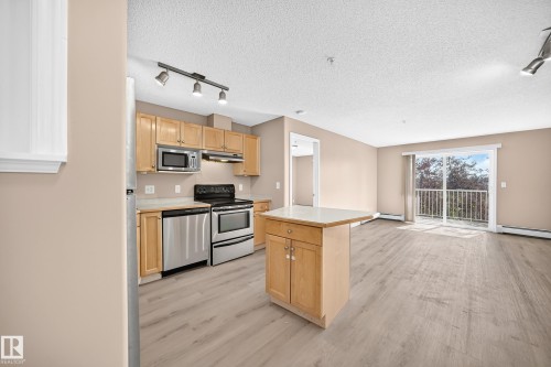 Kitchen and living area featuring wood-finish flooring, light wood cabinetry, stainless steel appliances, a kitchen island with a light countertop, and a sliding glass door to an outdoor balcony - #317 245 Edwards Dr Sw, Edmonton, AB - Indoor Photo Showing Kitchen