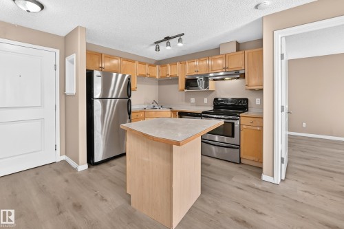 Modern kitchen featuring wood-finish flooring, stainless steel appliances, a corner sink, light wood cabinetry, and a central island with an extended countertop - #317 245 Edwards Dr Sw, Edmonton, AB - Indoor Photo Showing Kitchen With Stainless Steel Kitchen With Double Sink