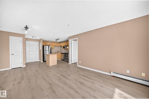 Open-concept living area featuring wood-finish flooring, a kitchen with light wood cabinetry, a stainless steel refrigerator, and a freestanding island - #317 245 Edwards Dr Sw, Edmonton, AB - Indoor