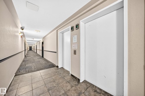Hallway featuring two elevators with white doors, tile flooring, and wall-mounted lighting fixtures - #317 245 Edwards Dr Sw, Edmonton, AB - Indoor Photo Showing Other Room