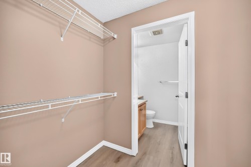 Walk-in closet with wire shelving system and wood-finish flooring, opening to a private bathroom featuring a wood-finish vanity with countertop and a toilet - #317 245 Edwards Dr Sw, Edmonton, AB - Indoor With Storage