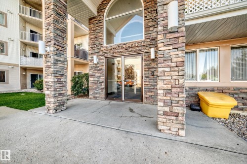 Building entrance featuring a stone facade, a large arched window above double glass doors, and concrete walkways - #317 245 Edwards Dr Sw, Edmonton, AB - Outdoor With Balcony With Facade
