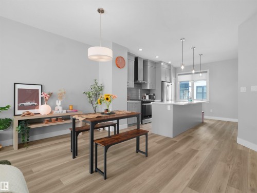 Open-concept living area featuring wood-finish flooring, a contemporary kitchen with light grey cabinetry, stainless steel appliances, and an island with pendant lighting - #81 6905 25 Av Sw, Edmonton, AB - Indoor
