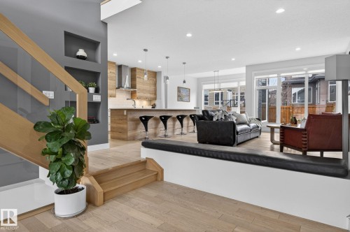 Open-concept living area featuring light wood-finish flooring, a contemporary staircase with glass railing, recessed wall shelving, a kitchen with wood-panel accents and stainless steel appliances, and large windows - 1313 Hainstock Way Sw, Edmonton, AB - Indoor